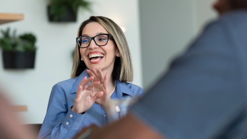 A woman in professional clothing and glasses laughs.