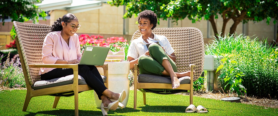 Two women sitting in chairs outside on a sunny day chatting and working on a laptop