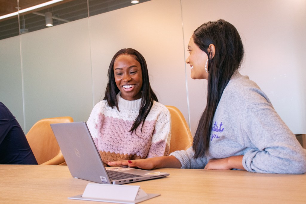 Two women smiling as they look at the same laptop together.
