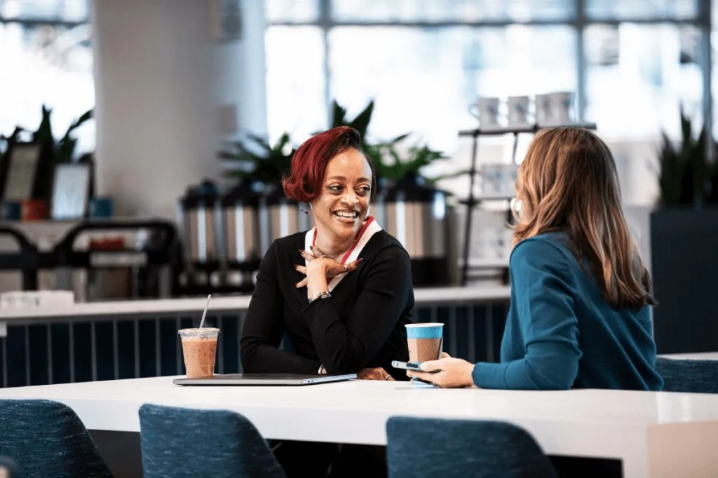 Two women sit at a table, talking over coffees.