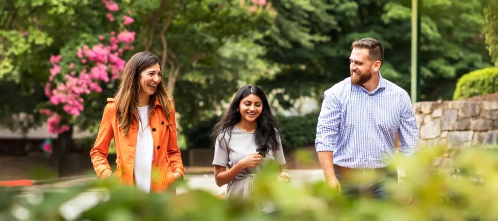 3 people walking outside, surrounded by foliage