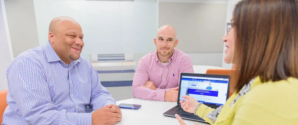 3 people sit and talk in a room with a white board and laptop.