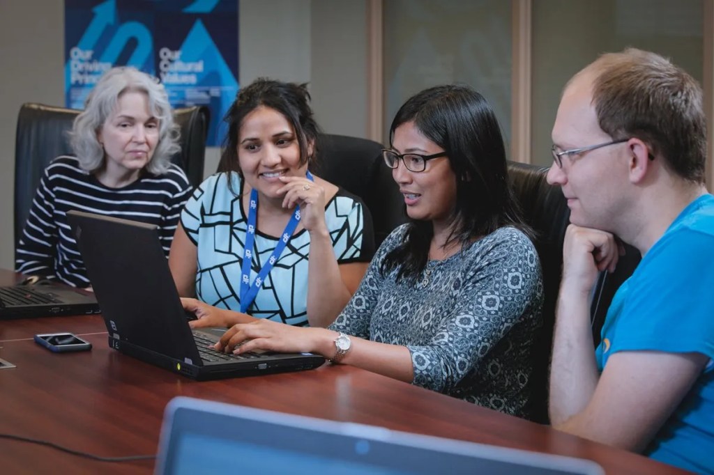 Four smiling colleagues huddle together and look at a laptop screen