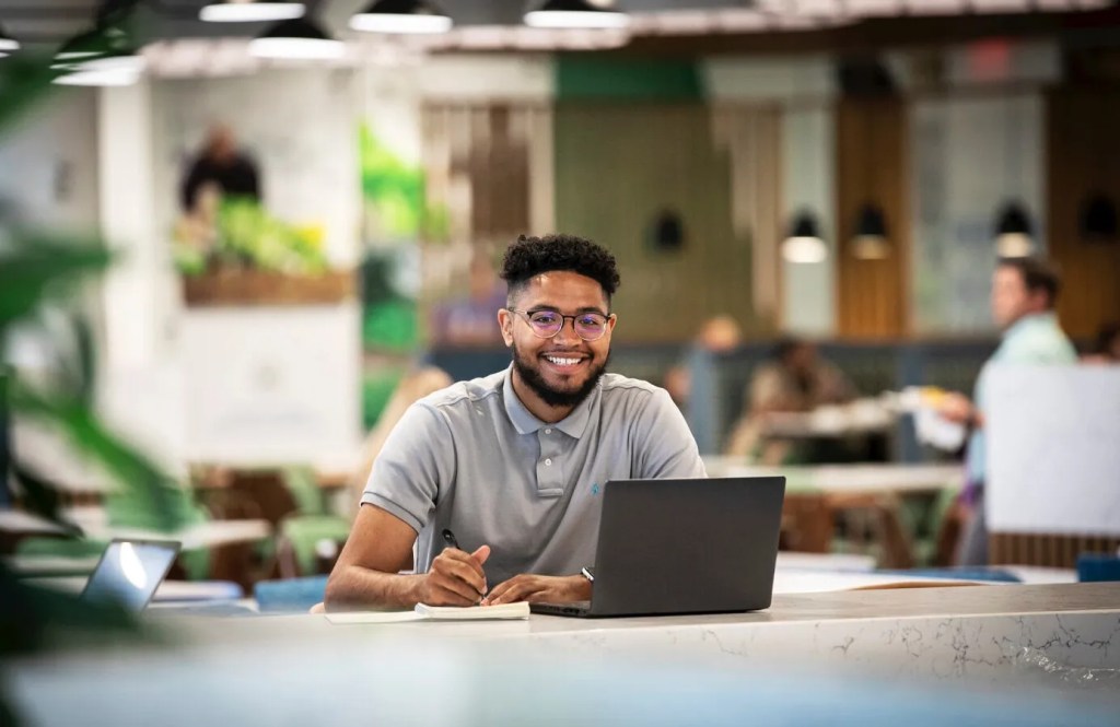 A smiling young man sits in an open concept working space with his laptop and notebook.