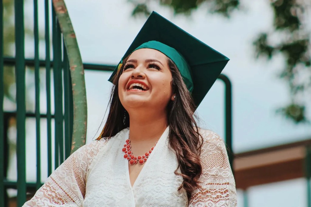 A young woman wearing a formal dress and graduation cap smiles and looks up at the sky. 