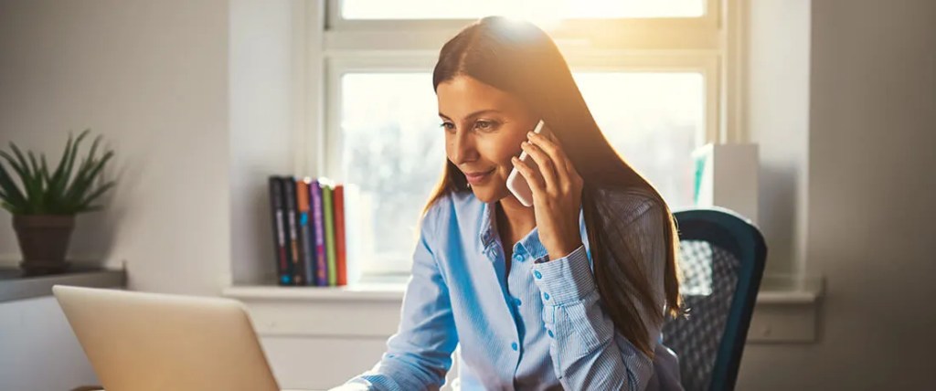Woman in a blue button up shirt on a cell phone call sitting at her desk.