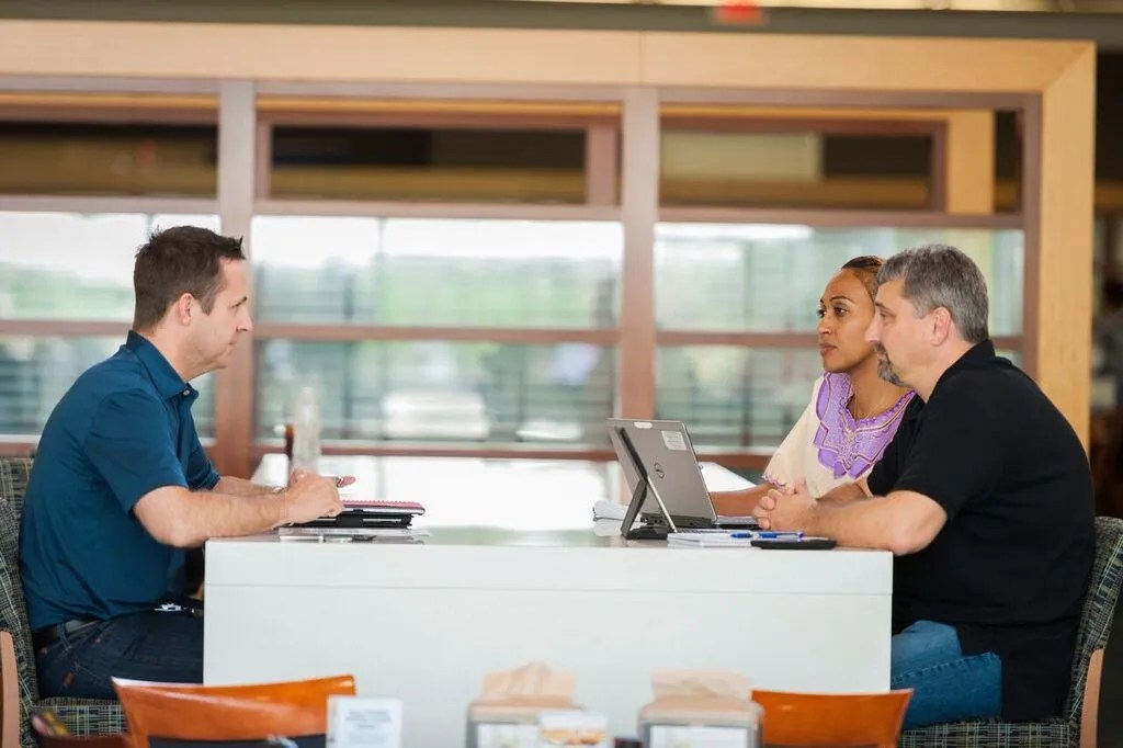 A man sits across a desk from another man and woman. They seem deep in discussion.
