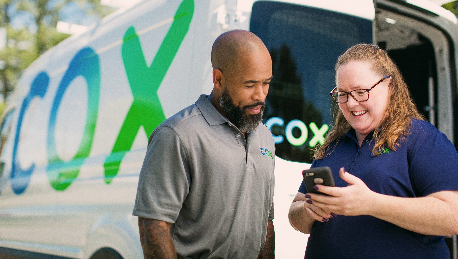 Two Cox employees reviewing and discussing what's seen on a phone in front of a Cox broadband service truck